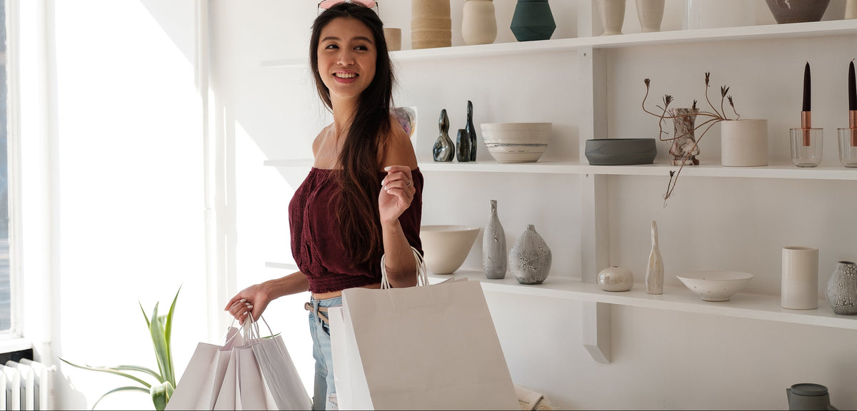 A woman smiling and holding a white shopping bag stands in a bright, modern room with white shelves displaying ceramic vases and bowls.